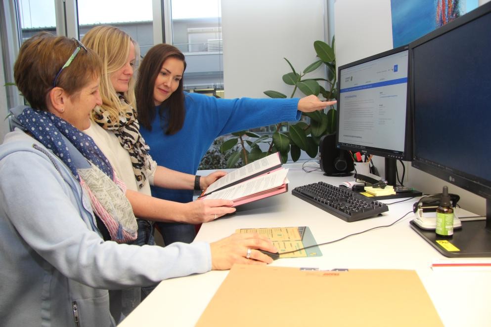 Drei Frauen stehen an einem Tisch. Eine der Frauen hält einen ausgedruckten Bauantrag in der Hand, eine andere zeigt auf einen Bildschirm, auf dem die Seite "Digitaler Bauantrag" aufgerufen ist. Die dritte Frau klickt mit einer Maus.