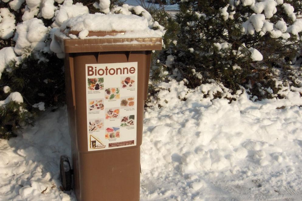 Auf dem Foto ist eine braune Biotonne zu sehen. Sie steht mitten im Schnee und ist teilweise auch zugeschneit.